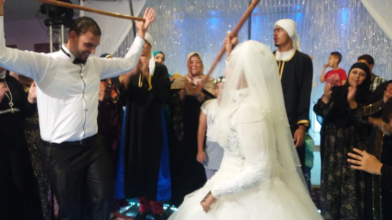 عريس وعروس يرقصان بعصا التحطيب في فرح مصري، والمدعوون يصفّقون حولهما
