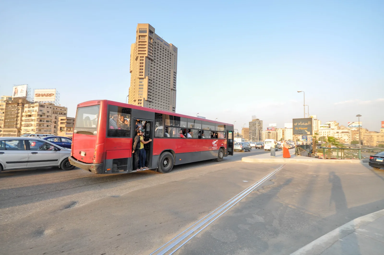أتوبيس نقل عام أحمر يسير على كوبري السادس من أكتوبر وراكبان يتعلقان بباب مفتوح، والسيارات حوله