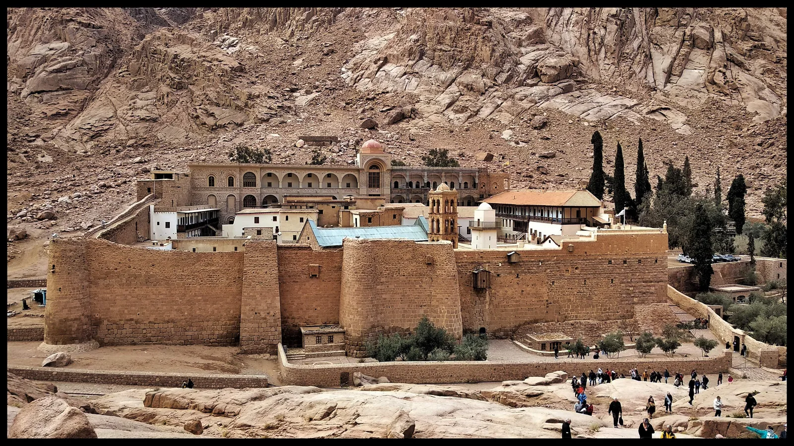 دير سانت كاترين بأسواره الحجرية المحصّنة عند سفح جبال جنوب سيناء، وزوار يسيرون أمامه